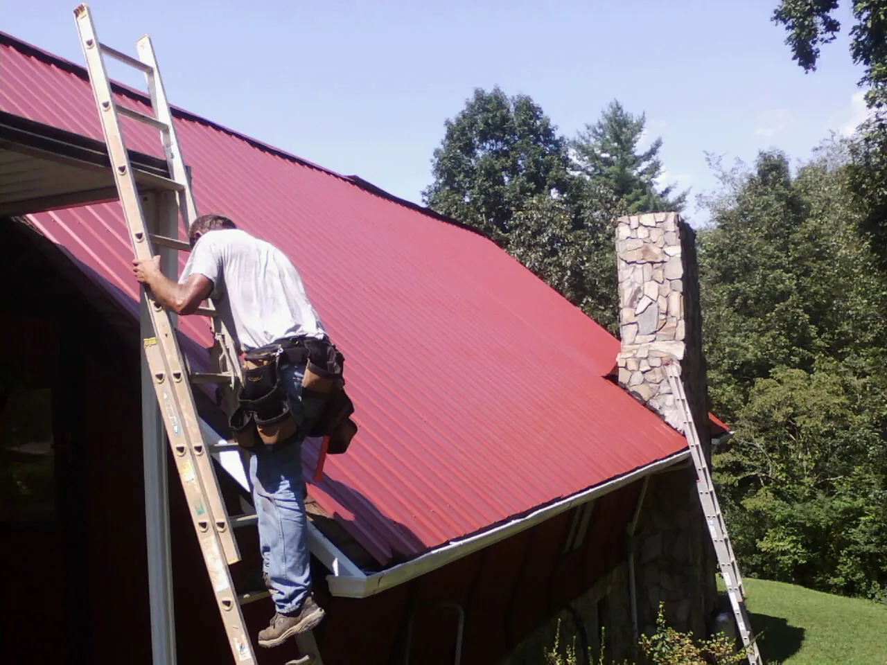 Roofer on a ladder beside a red metal roof and stone chimney — Soffit & Fascia Repair by Roofing Brothers in Brook Park